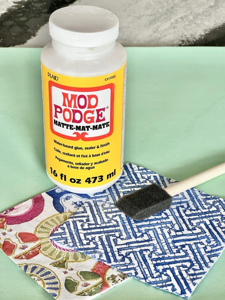 Supplies for DIY decoupage coasters that include Mod Podge, paper napkins, and a foam brush.