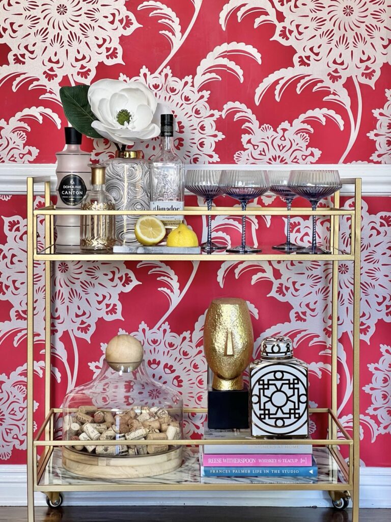 A gold bar cart filled with glamorous decorating ideas of glass vases, crystal drink glasses, a gold head bust, and. single magnolia bloom.