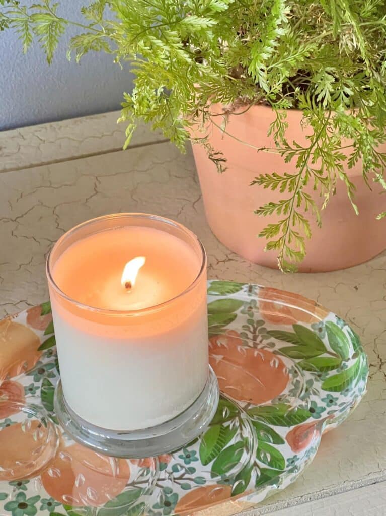 A burning candle on a orange and green tissue paper decoupaged glass snack tray.