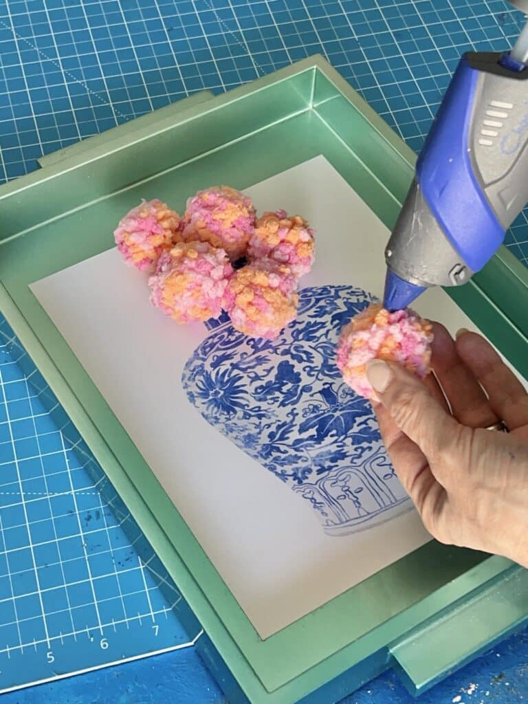 Using a hot glue gun to attach the poms to the art print for yarn wall decor diy.