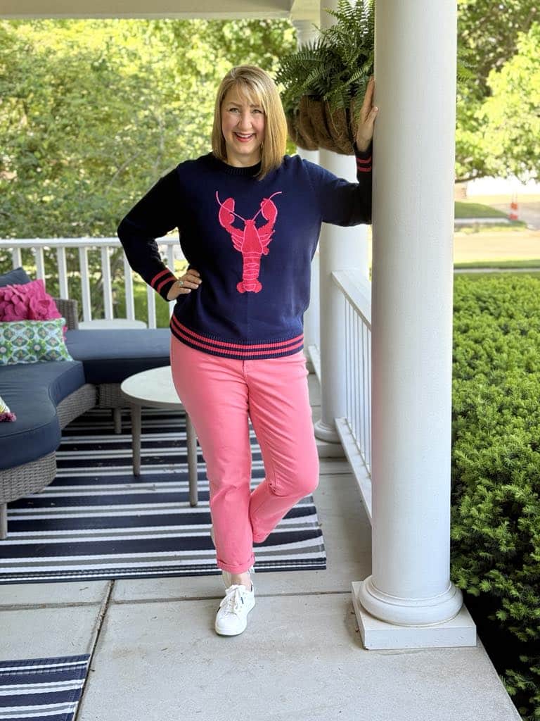 how to dress preppy: Woman wearing a blue sweater with a pink embroidered lobster and salmon colored jeans.