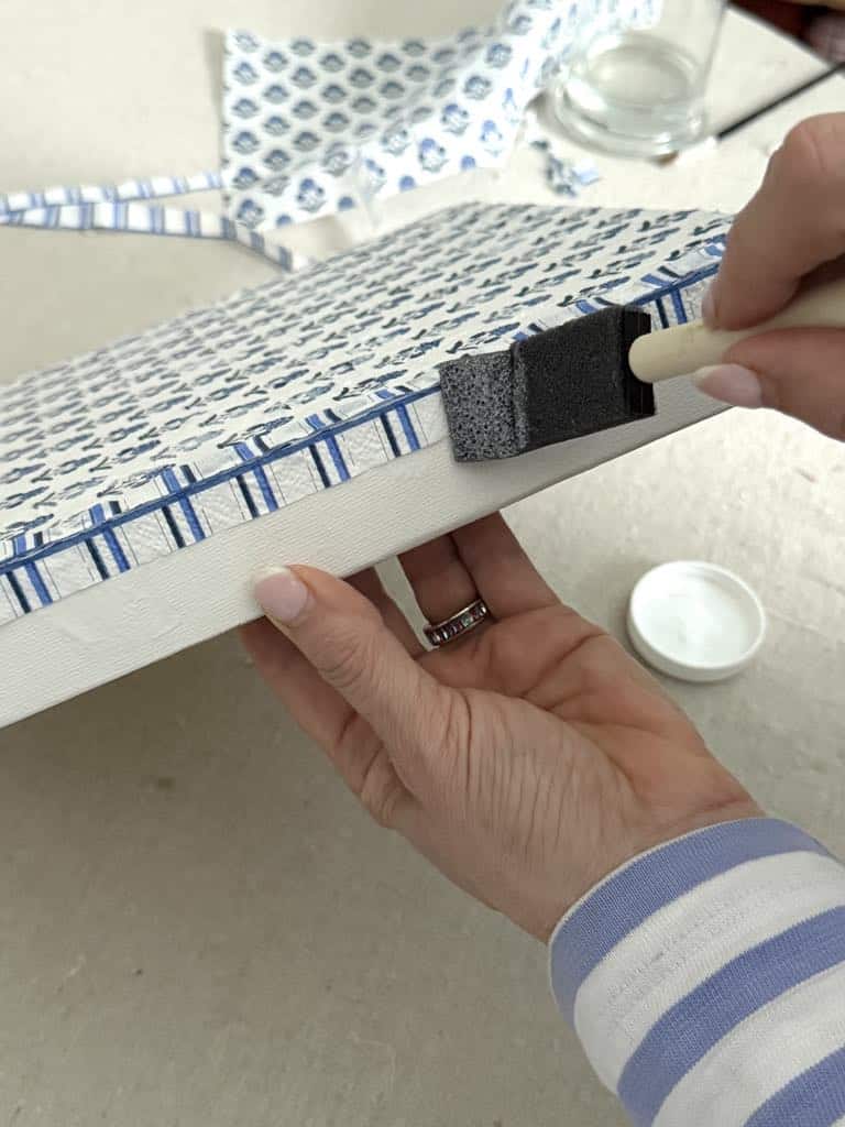 Brushing decoupage glue over a paper napkin on the side of a wrapped canvas.