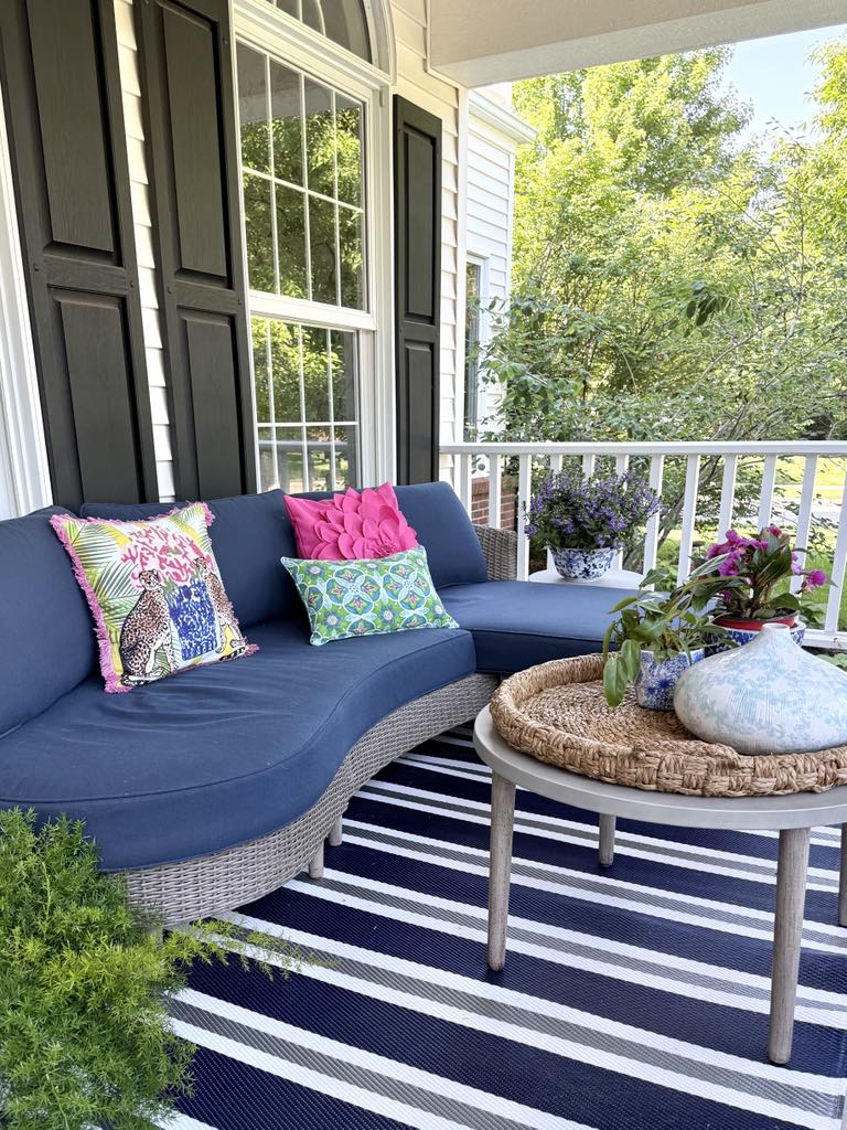 Outdoor sofa decorated with summer pillows and sitting on a front porch. An outdoor coffee table is styled with plants and pots and a ceramic vase.