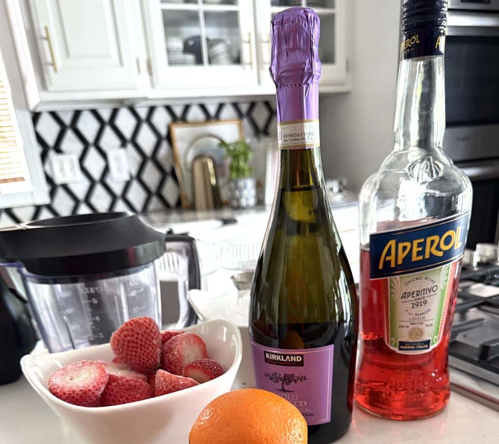 A bottle of aperol, prosecco, and a bowl of frozen strawberries.