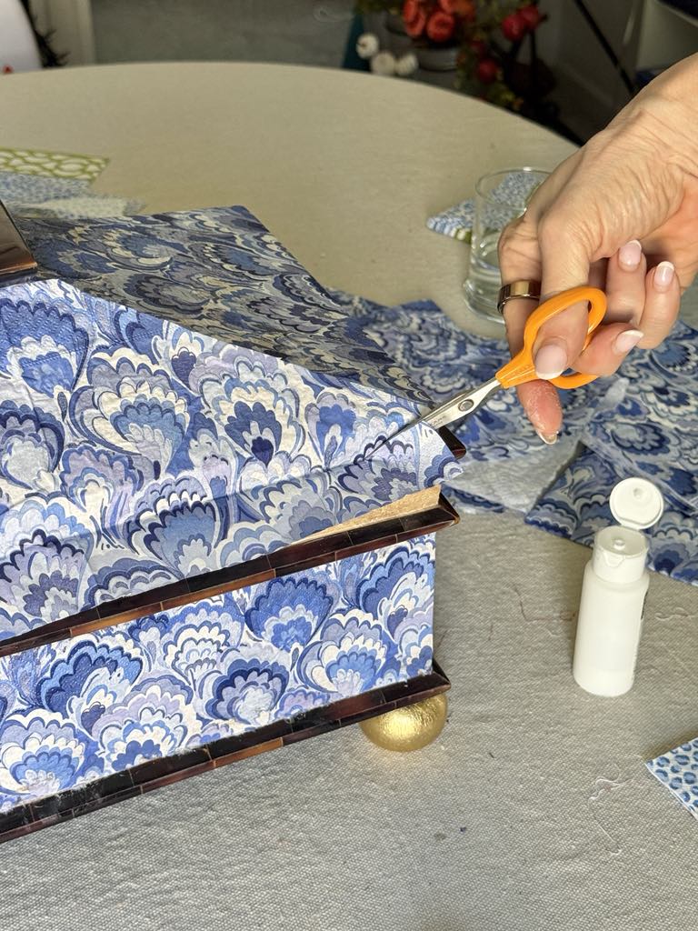 Trimming the excess napkin from a decoupaged box with scissors.