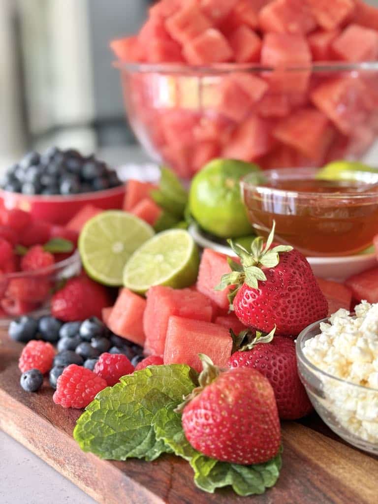 Strawberries, blueberries, raspberries, watermelong, limes, feta cheese, and lime on a charcuterie board.
