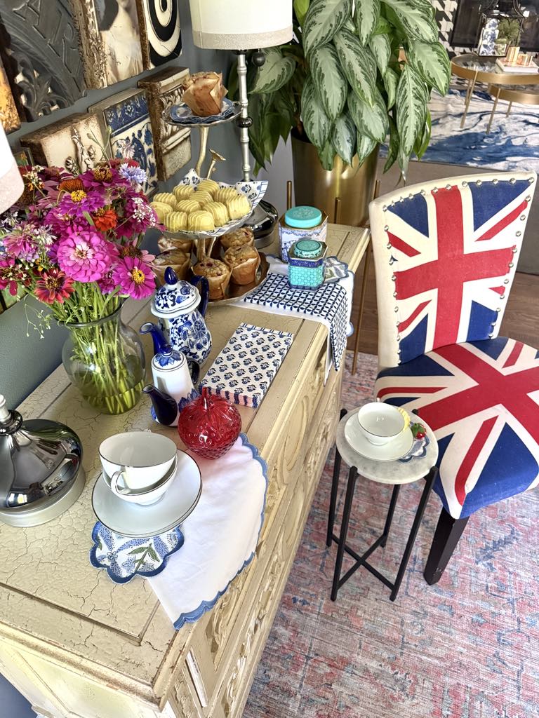 A tea party tablescape with pink flowers, blue and white linens, white china, and a Union Jack upholstered chair.