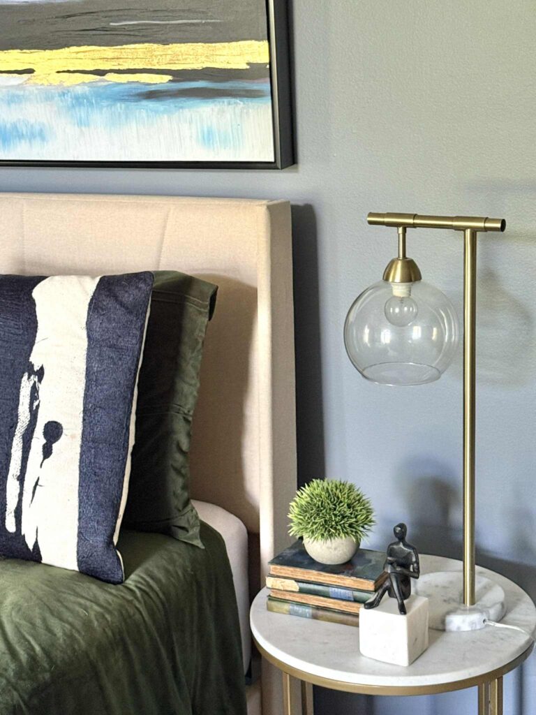 A bedroom nightstand styled with a glass lamp, a black and white sculpture, and a green plant.