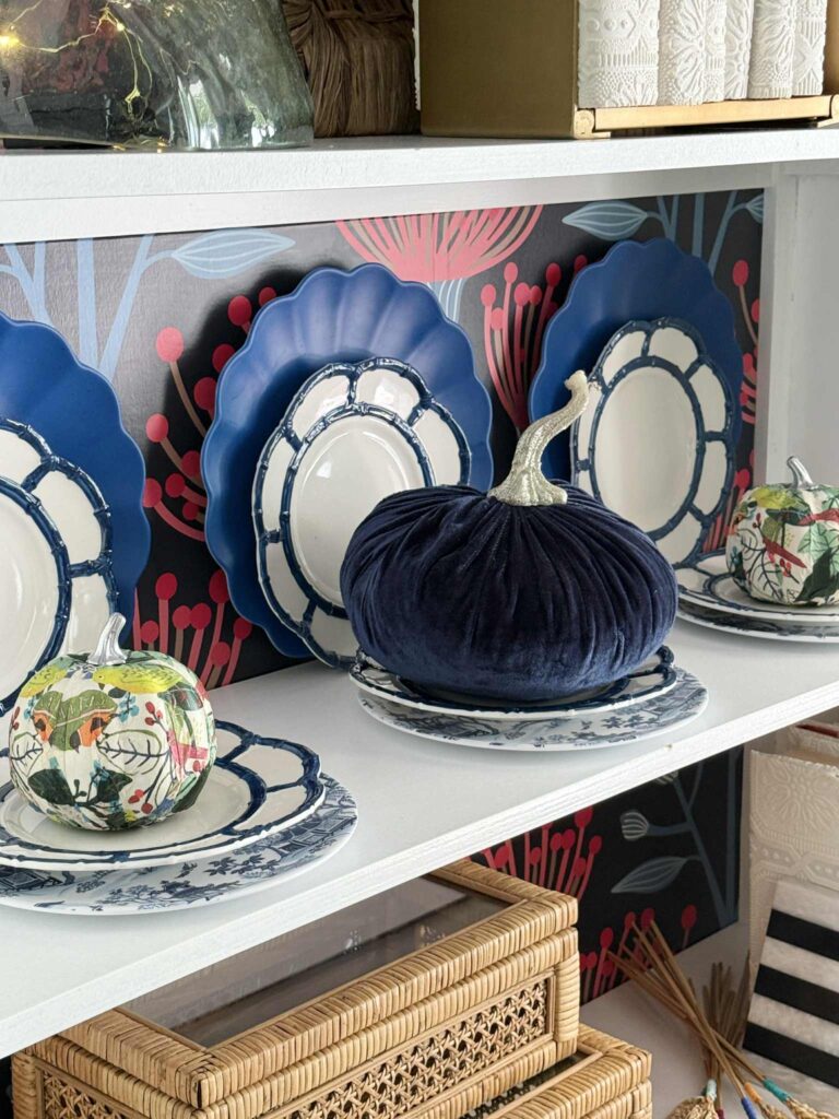 A bookshelf holding stacked plates and colorful pumpkins.