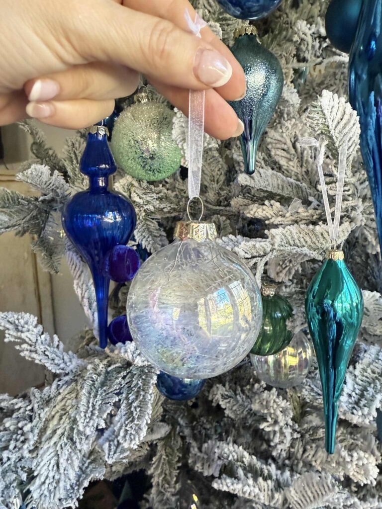A woman's hand holding a clear ornament in front of a flocked tree decorated for Christmas.