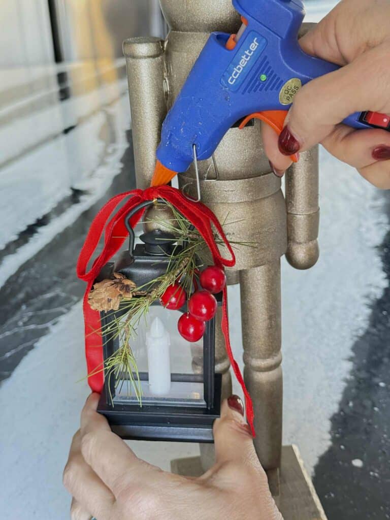 Using hot glue gun to attach a tiny lantern embellished with greenery and berries to the wooden nutcracker hand.