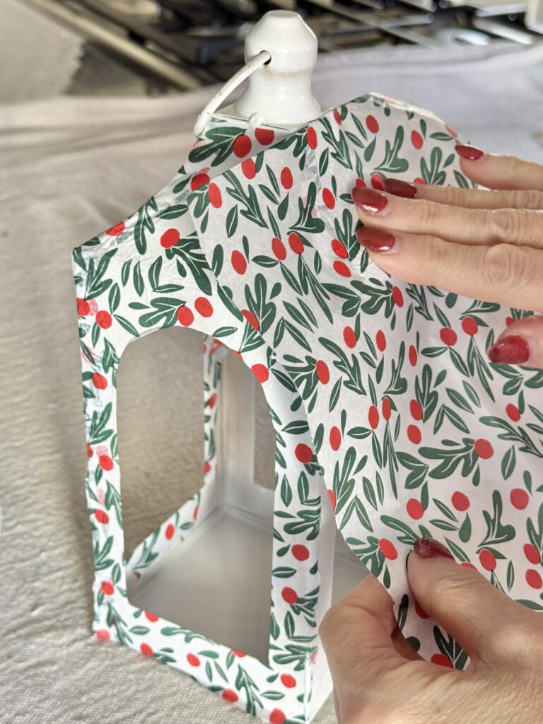 Pressing Christmas themed tissue paper onto decoupage glued surface of a metal lantern.