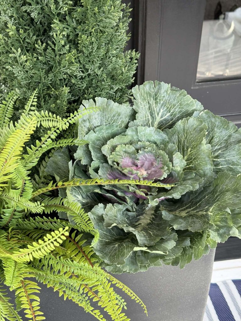 A porch planter filled with a faux cabbage, fern, and evergreen shrub.