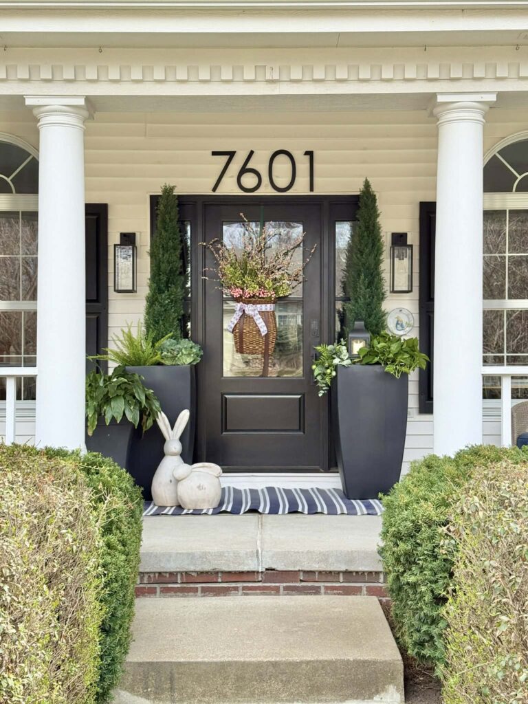 A front porch with a black door, black planters, layers of greenery, and a hanging basket on the front door.