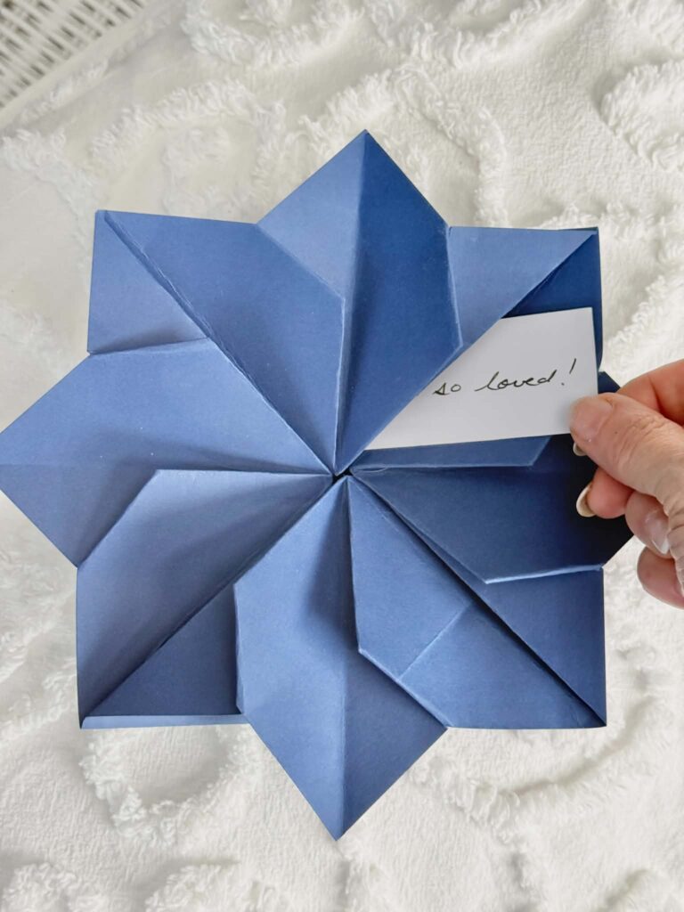 A woman's hand pulling a handwritten note from inside a flower origami envelope.