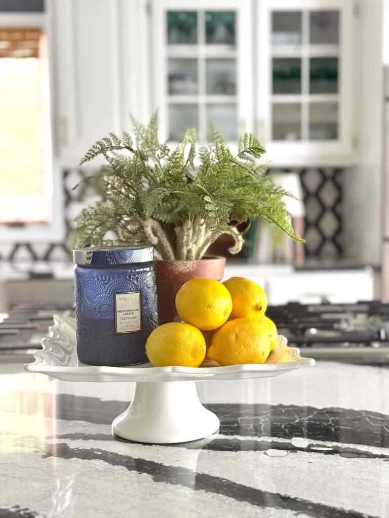 A cake stand holding fresh lemons, a jar candle, and a green plant.