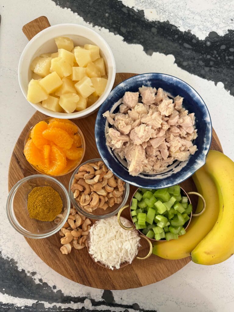 Chicken, pineapple, mandarin oranges, celery, curry powder, flaked coconut, and bananas on a wood board.