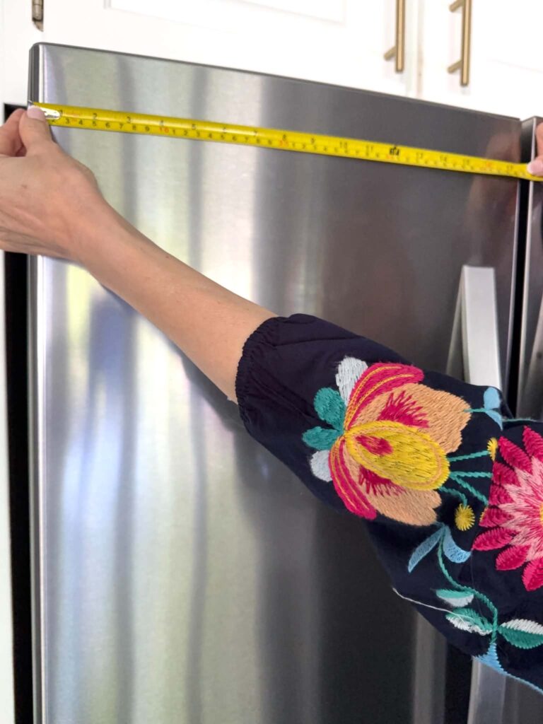 A woman measuring the top of a refrigerator door before applying peel and stick wallpaper.