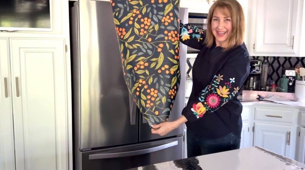 A woman in font of a refrigerator displaying a roll of wallpaper with an orange vine pattern.